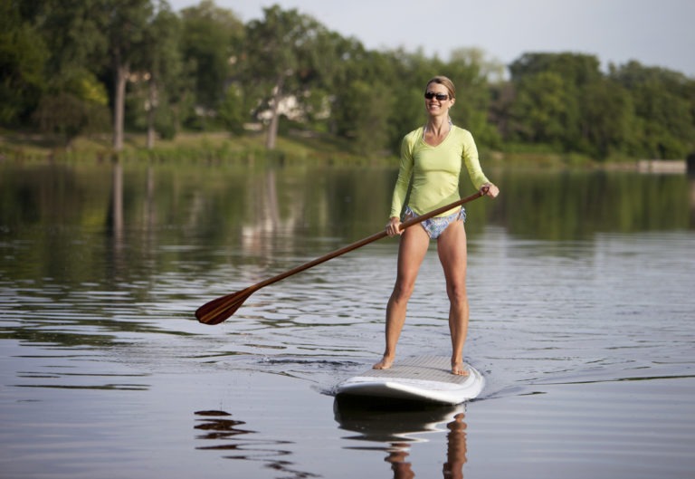 Woman standing on a paddleboard on a calm Colorado lake, wearing a bright green rash guard and paddling on a sunny day